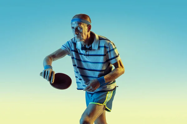 Старший мужчина играет в настольный теннис в спортивной форме на градиентном фоне и неоновом свете — стоковое фото