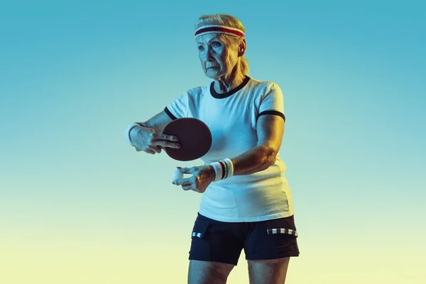 Mulher sênior treinando em tênis de mesa em sportwear em fundo gradiente em luz de néon — Fotografia de Stock