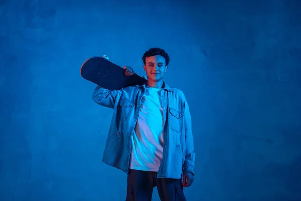 Kaukasischer junger Skateboarder posiert auf dunklem neonbeleuchtetem Hintergrund — Stockfoto