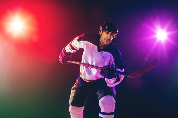 Pemain hoki pria dengan tongkat di lapangan es dan latar belakang berwarna neon gelap — Stok Foto