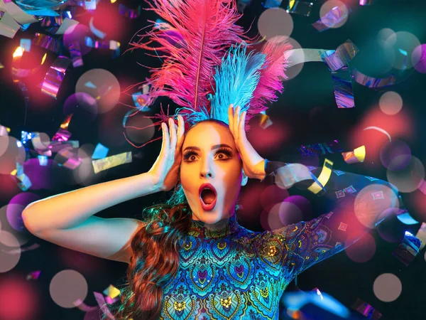 Beautiful young woman in carnival and masquerade costume in colorful neon lights on black background in flying confetti