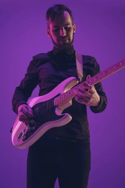 Jovem músico caucasiano tocando guitarra em luz de néon em fundo roxo — Fotografia de Stock