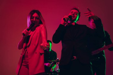 Genç beyaz müzisyenler, şarkıcılar kırmızı stüdyo arka planında neon ışıkta şarkı söylüyorlar.