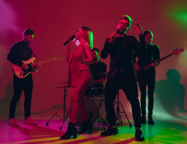 Kafkasyalı genç müzisyenler, kırmızı stüdyo arka planında neon ışıkta performans sergiliyorlar.
