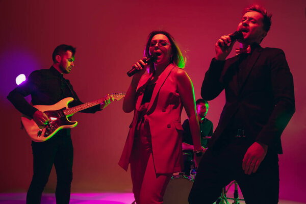 Young caucasian musicians, band performing in neon light on red studio background