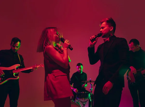 Young caucasian musicians, band performing in neon light on red studio background