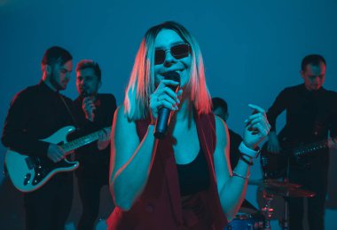 Kafkasyalı genç müzisyenler, mavi stüdyo arka planında neon ışığı altında şarkı söyleyen orkestra, öndeki şarkıcı.