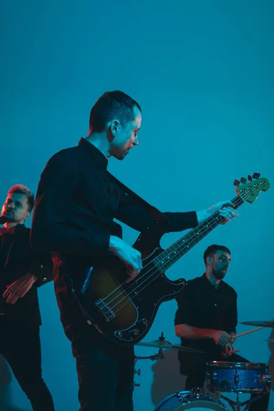 Junge kaukasische Musiker, Band spielt im Neonlicht auf blauem Studiohintergrund, Gitarrist vorn — Stockfoto