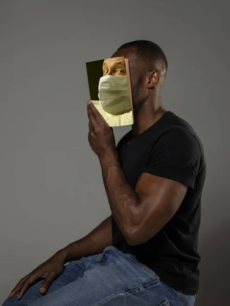 Auf Nummer sicher gehen und lesen, um jemand anderer zu werden - Mann verdeckt Gesicht mit Buch in Gesichtsmaske, während er auf grauem Hintergrund liest — Stockfoto