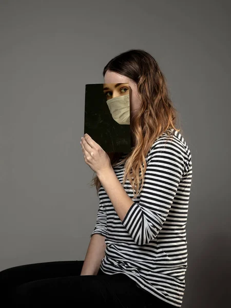 Να είστε ασφαλείς και διαβάστε για να γίνετε κάποιος άλλος - γυναίκα που καλύπτει το πρόσωπο με το βιβλίο στη μάσκα προσώπου ενώ διαβάζετε στο γκρίζο υπόβαθρο — Φωτογραφία Αρχείου