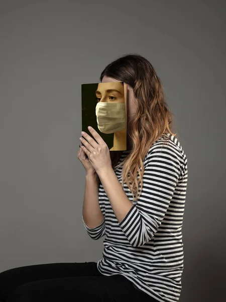 Sicher sein und lesen, um jemand anderes zu werden - Frau verdeckt Gesicht mit Buch in Gesichtsmaske, während sie auf grauem Hintergrund liest — Stockfoto