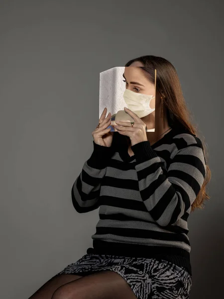Bądź bezpieczny i czytaj, aby stać się kimś innym - kobieta zakrywa twarz książką w masce twarzy czytając na szarym tle — Zdjęcie stockowe