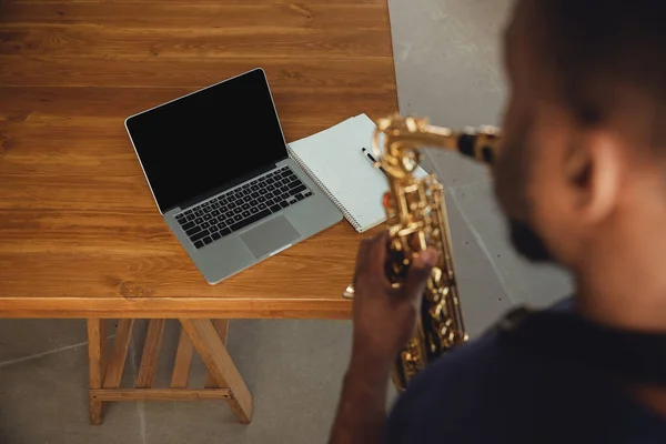 African-american musician playing saxophone during online concert at home isolated and quarantined. Blank laptop screen with copyspace.