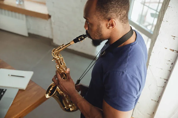 African-american musician playing saxophone during online concert at home isolated and quarantined, impressive improvising