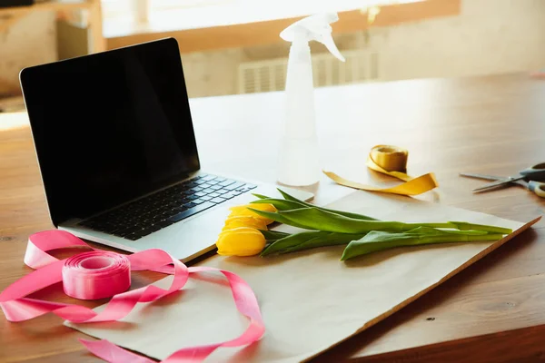 Флорист за работой: стол с тюльпанами и нарциссами, ленты и покрывающая бумага против ноутбука, подготовленный для мастерской — стоковое фото