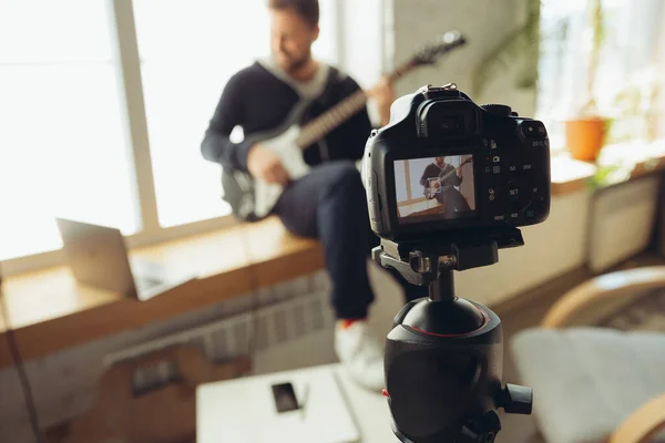 Músico caucásico tocando la guitarra durante el concierto en casa aislado y en cuarentena, improvisación alegre, enfoque en la cámara — Foto de Stock