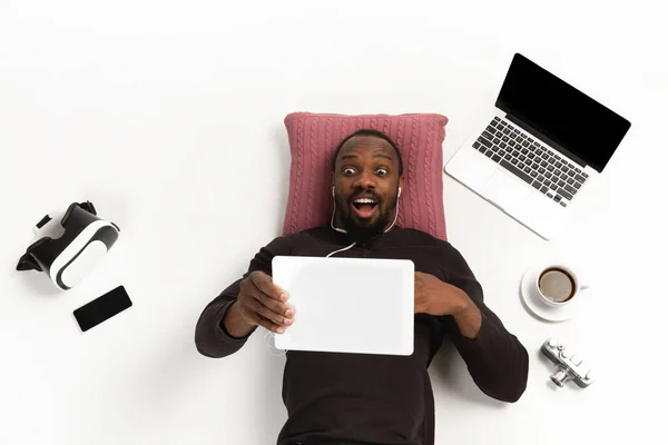 Homem afro-americano emocional usando tablet cercado por gadgets isolados em fundo de estúdio branco, tecnologias que conectam as pessoas. Chocado, assustado — Fotografia de Stock