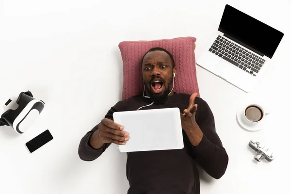 Emotionale afrikanisch-amerikanische Mann mit Tablet umgeben von Gadgets isoliert auf weißem Studiohintergrund, Technologien. Wütend gefragt — Stockfoto