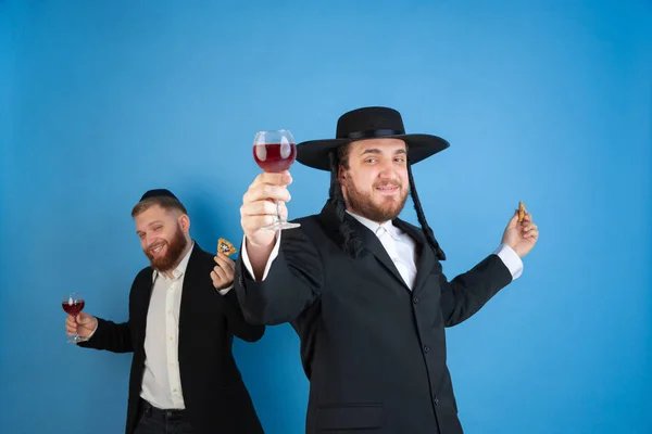 Retrato de um jovem judeu ortodoxo isolado no fundo do estúdio azul, encontrando a Páscoa, comendo orelhas Amans com vinho — Fotografia de Stock