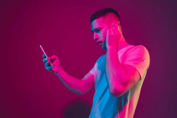Vit mans porträtt isolerad på rosa-lila studio bakgrund i neon ljus, med smartphone och hörlurar — Stockfoto