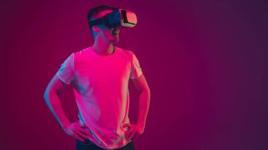 Beyaz adam portresi pembe-mor stüdyo arka planında, neon ışıkta izole edilmiş, VR-kulaklıkla oynuyor.