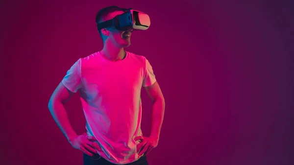 Biały portret mężczyzny odizolowany na różowo-fioletowym tle studia w neonowym świetle, bawiący się słuchawkami VR — Zdjęcie stockowe