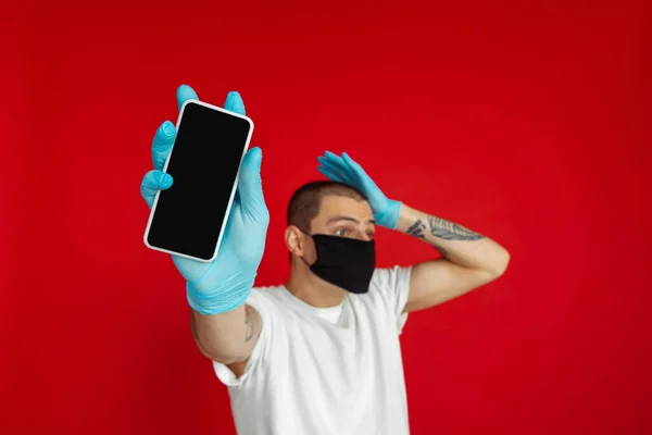 Καυκάσιος νεαρός άνδρας με προστατευτική μάσκα προσώπου και ιατρικά γάντια σε κόκκινο φόντο στούντιο - που δείχνει λευκό οθόνη τηλέφωνα — Φωτογραφία Αρχείου