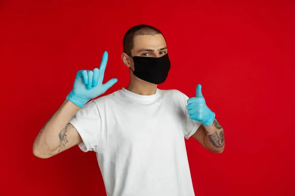 Hombre joven caucásico con mascarilla protectora y guantes médicos en el fondo rojo del estudio - señalando y mostrando el pulgar hacia arriba — Foto de Stock