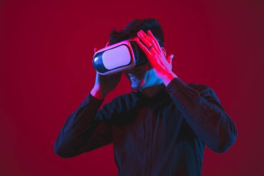 Beyaz adamın portresi kırmızı stüdyo arka planında, mor-mavi neon ışıkla izole edilmiş, duygusal ve güzel, VR kulaklık takıyor.