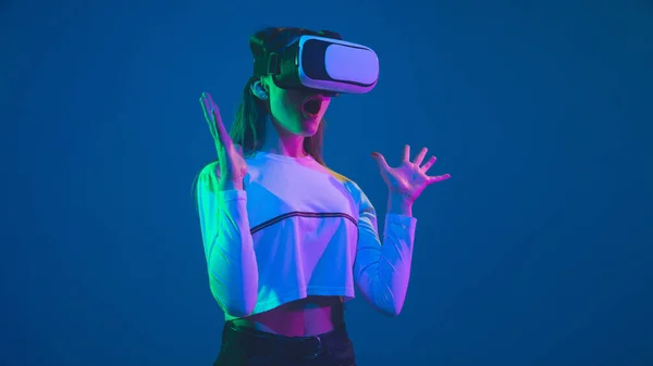 Retrato de mujer caucásica aislado sobre fondo de estudio azul en luz de neón rosa-verde, elegante y hermoso, jugando con auriculares VR —  Fotos de Stock