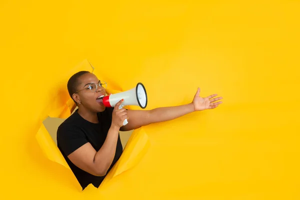 Jovem alegre posa em papel amarelo rasgado buraco fundo, emocional e expressivo, gritando e chamando com alto-falante — Fotografia de Stock