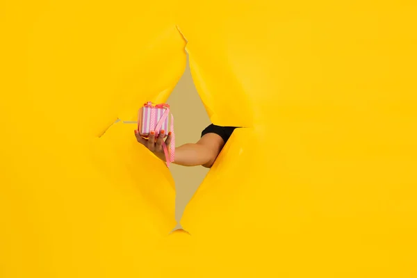 Mão feminina dando presente em rasgado papel amarelo buraco fundo, celebração — Fotografia de Stock
