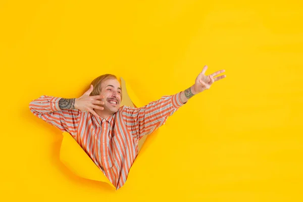 Χαρούμενος νεαρός άνδρας θέτει σε σχισμένο κίτρινο φόντο τρύπα χαρτί, συναισθηματική και εκφραστική — Φωτογραφία Αρχείου
