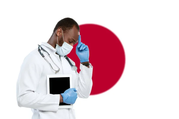 Молодой врач со стетоскопом и маской для лица молится за Бога с японским национальным флагом на заднем плане — стоковое фото