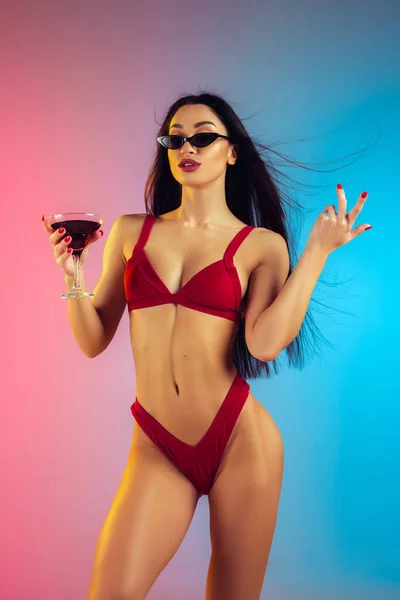 Fashion portrait of young fit and sportive woman with cocktail in stylish red luxury swimwear on gradient background. Perfect body ready for summertime. — Stock Photo, Image