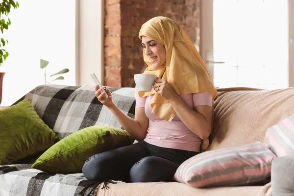 Красивая молодая мусульманка дома во время карантина и самоизоляции, используя смартфон для селфи или видео, онлайн-уроков, шоппинга — стоковое фото