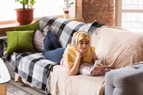 Красивая молодая мусульманка дома во время карантина и самоизоляции, используя наушники, слушая музыку, смотря кино, сериалы, наслаждаясь — стоковое фото
