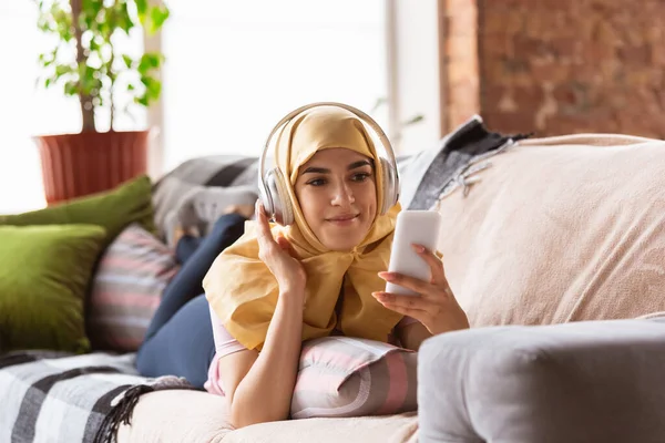 Красивая молодая мусульманка дома во время карантина и самоизоляции, используя наушники, слушая музыку, смотря кино, сериалы, наслаждаясь — стоковое фото