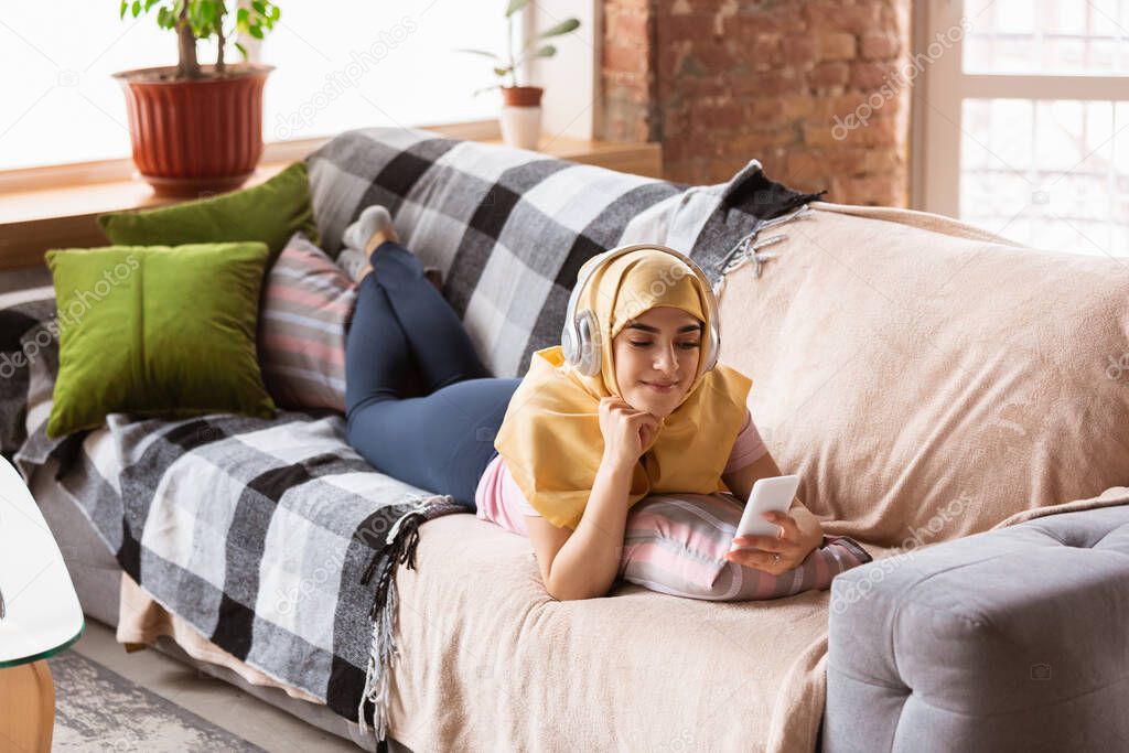 A pretty young muslim woman at home during quarantine and self-insulation, using headphones, listen to music, watching cinema, serials, enjoying