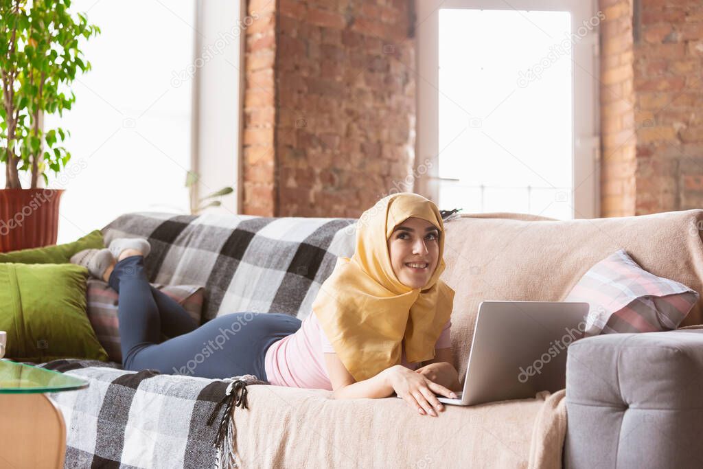 A pretty young muslim woman at home during quarantine and self-insulation, using laptop, listen to music, watching cinema, serials, shopping, studying