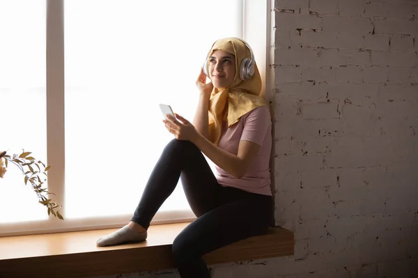 Красивая молодая мусульманка дома во время карантина и самоизоляции, используя смартфон для селфи или видео, онлайн-уроков, шоппинга — стоковое фото