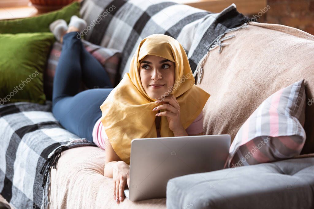 A pretty young muslim woman at home during quarantine and self-insulation, using laptop, listen to music, watching cinema, serials, shopping, studying