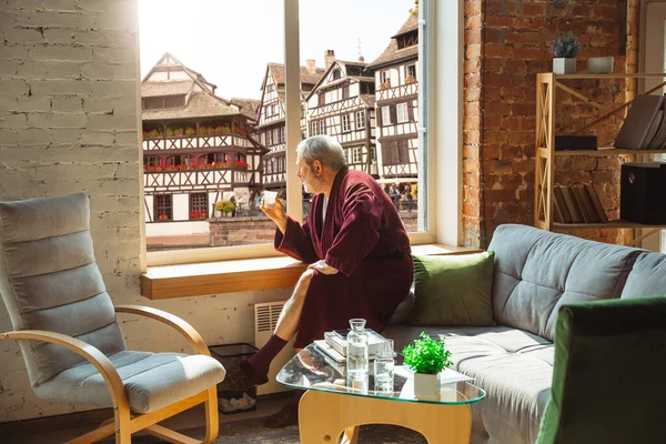 Dojrzały starszy mężczyzna podczas kwarantanny, zdając sobie sprawę, jak ważny jest pobyt w domu podczas epidemii wirusa, patrząc przez okno na dzień miasta we Francji — Zdjęcie stockowe