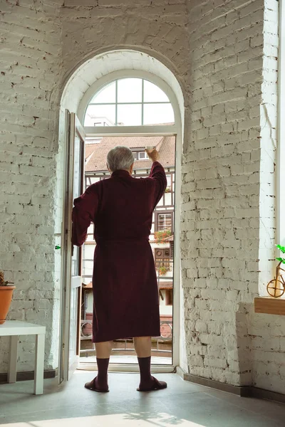 Hombre mayor maduro durante la cuarentena, dándose cuenta de la importancia de permanecer en casa durante el brote de virus, mirando a través de la ventana en la ciudad de día en Francia — Foto de Stock