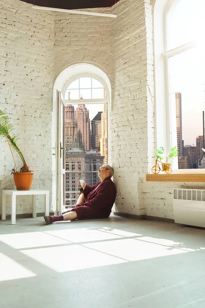 Homem idoso maduro durante a quarentena, percebendo o quão importante é ficar em casa durante o surto do vírus, olhando pela janela do dia Nova York — Fotografia de Stock