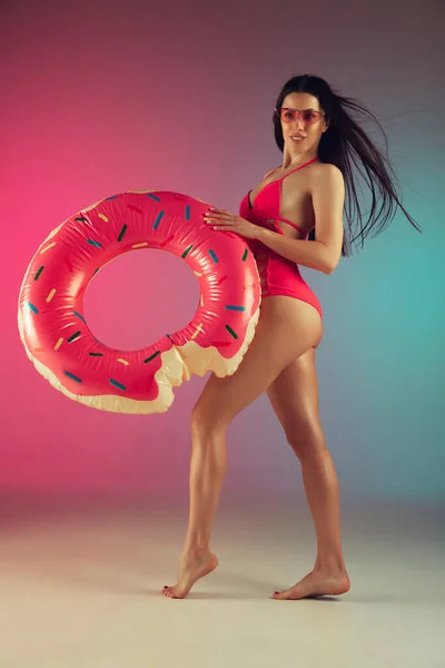 Retrato de moda de mulher jovem em forma e esportiva em trajes de banho de luxo rosa elegante com donut de borracha no fundo gradiente. Corpo perfeito pronto para o verão . — Fotografia de Stock