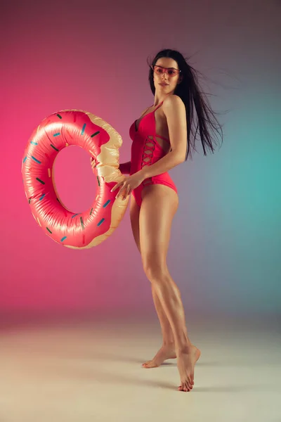 Retrato de moda de mulher jovem em forma e esportiva em trajes de banho de luxo rosa elegante com donut de borracha no fundo gradiente. Corpo perfeito pronto para o verão . — Fotografia de Stock