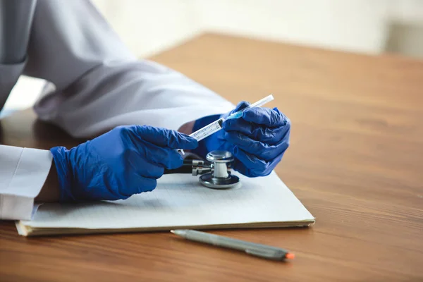 Gros plan des mains des médecins portant des gants de protection bleus avec stéthoscope et seringue sur fond de table en bois — Photo