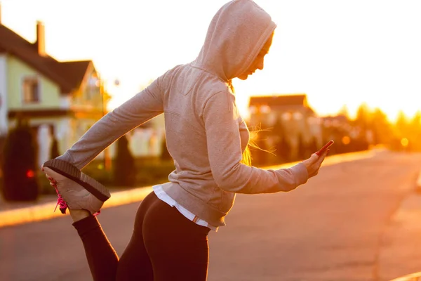 Jonge vrouwelijke loper, atleet is stretching voordat joggen in de stad straat in de zon. Mooie Kaukasische vrouw training, luisteren naar muziek — Stockfoto