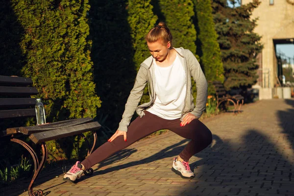 Joven corredora, atleta se está estirando antes de correr en la calle de la ciudad bajo el sol. Hermosa mujer caucásica entrenando, escuchando música — Foto de Stock